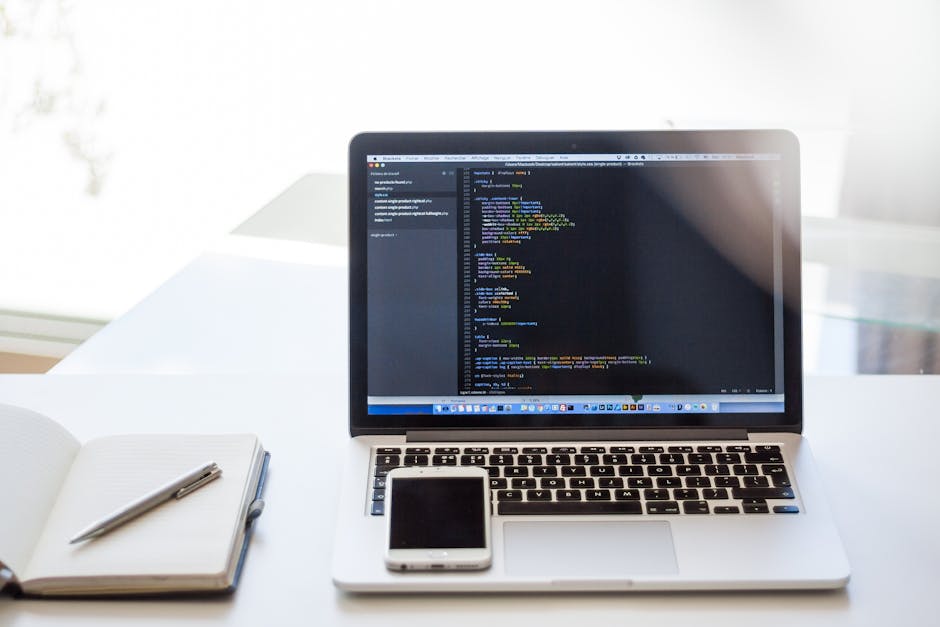 A close-up view of a modern workspace featuring a silver laptop on a white desk, displaying lines of colorful code on its screen, with a black smartphone positioned in front of the laptop. To the left